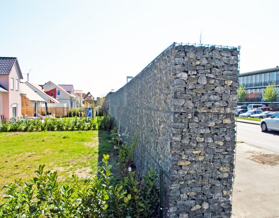 Hohe, graue Gabionenwand mit groben Steinen trennt ein Wohnquartier mit Gärten von einer Strasse mit parkenden Autos.