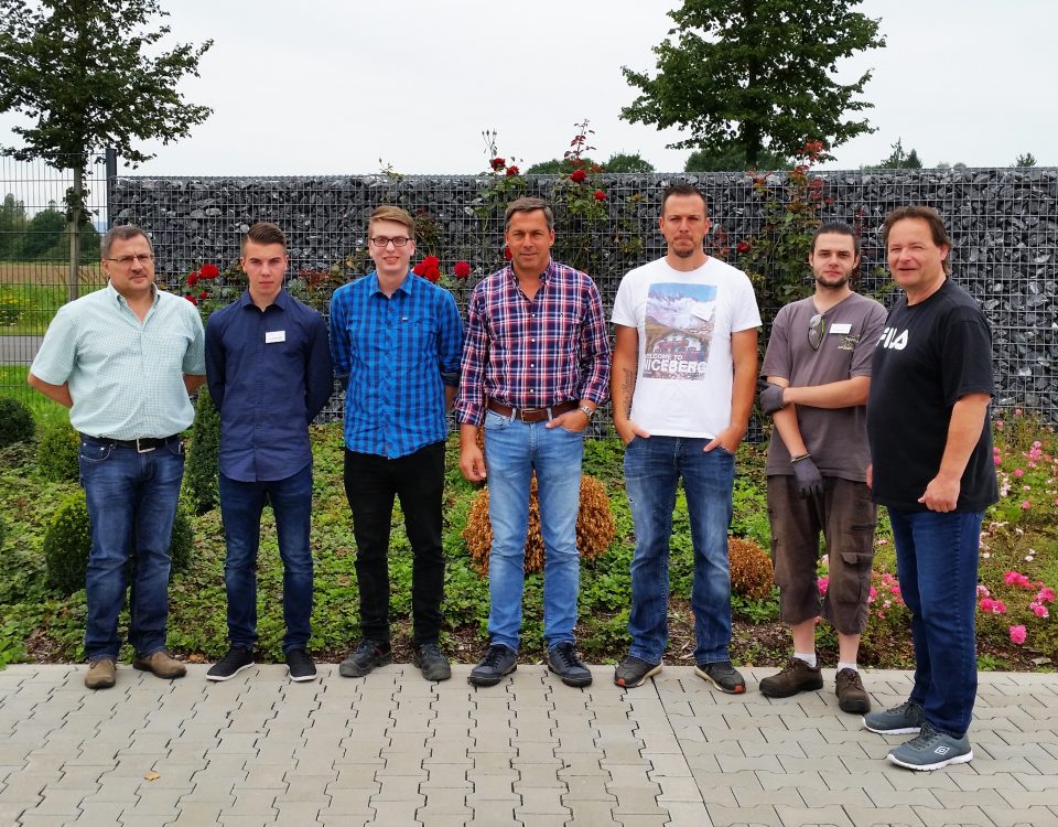 Gruppe von sieben Personen vor einer bepflanzten Gabionenwand in einem Aussenbereich.