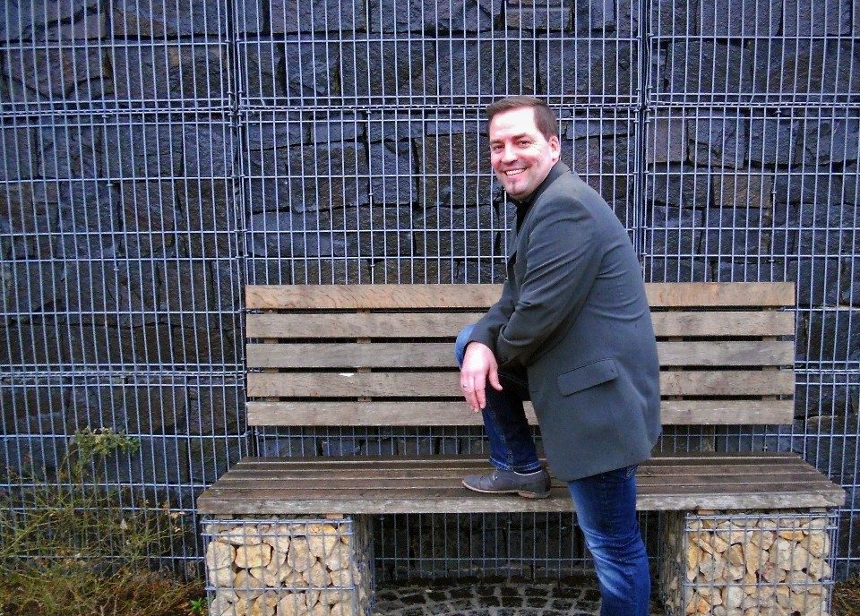 Ein Mann in Jeans und grauem Sakko sitzt lässig auf einer Holzbank, deren Unterkonstruktion aus Gabionen besteht. Hinter ihm steht eine hohe Gabionenwand aus dunklem Stein, die das gesamte Hintergrundbild ausfüllt.