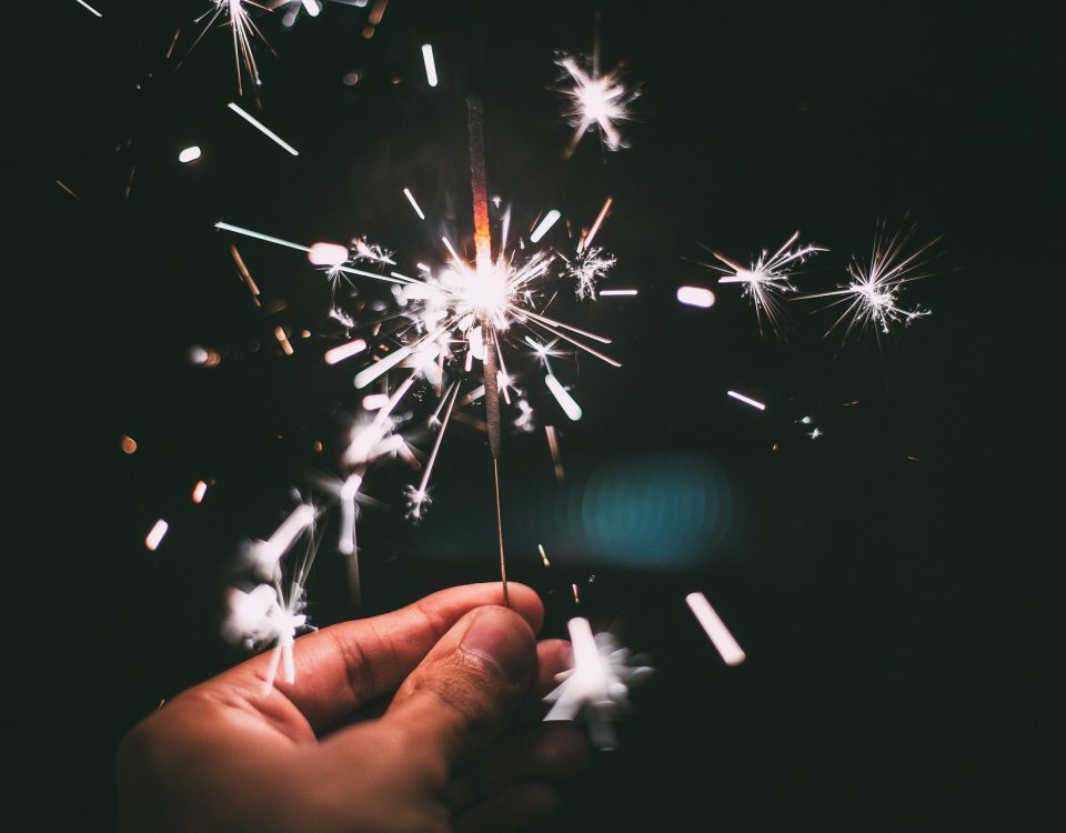 Hand hält eine brennende Wunderkerze vor dunklem Hintergrund mit fliegenden Funken.