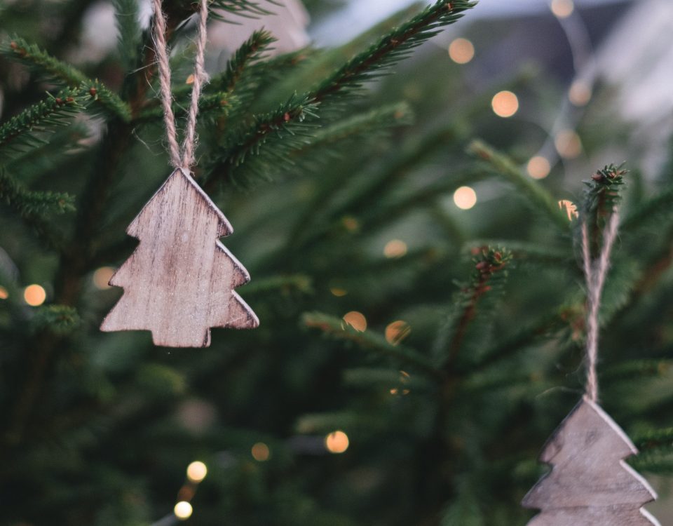 Holzanhänger in Tannenbaumform hängen an den Zweigen eines geschmückten Weihnachtsbaums, im Hintergrund warme Lichter in sanfter Unschärfe.